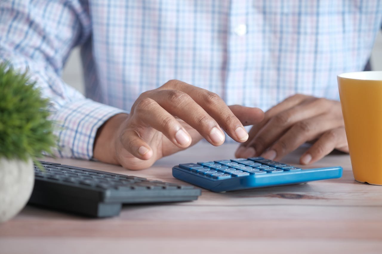 Home A person using a blue calculator on a wooden desk with a keyboard and plant nearby.